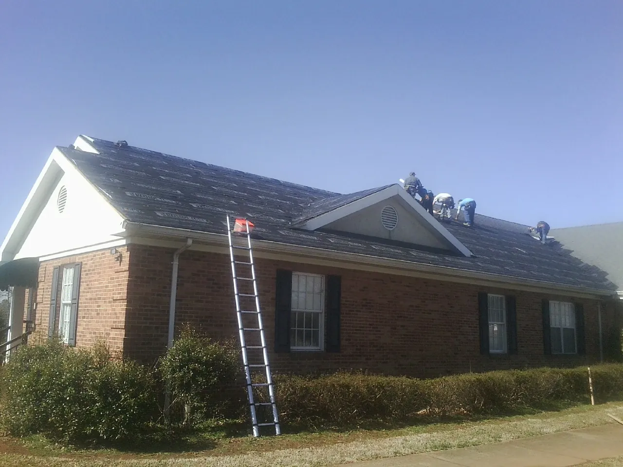 Crew tearing shingles off a brick ranch roof with a ladder set — Metal Roof Repair work by our crew in Ramsey