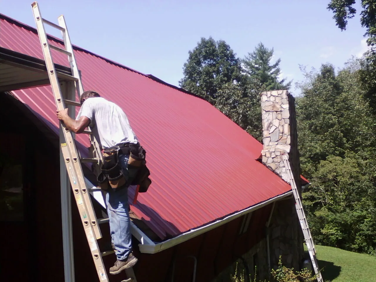 Roofer on a ladder beside a red metal roof and stone chimney in Ramsey